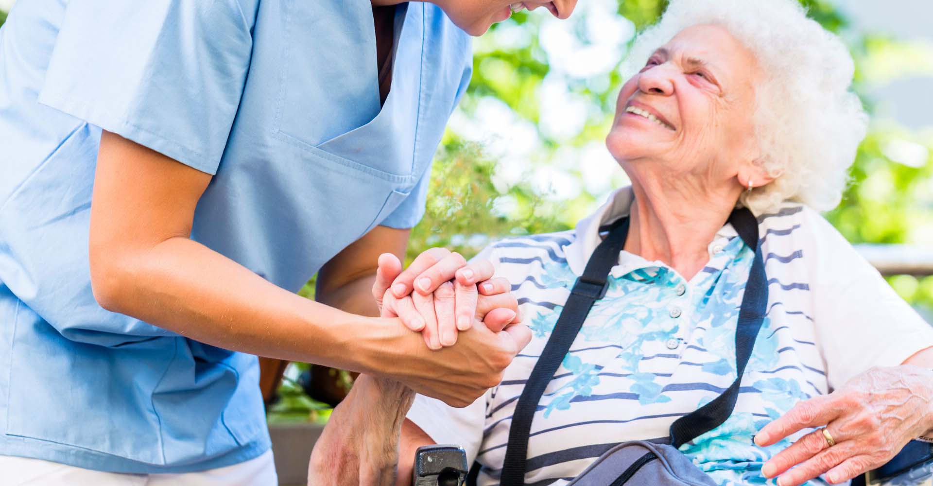 picture of an elderly woman sitting on a wheelchair. And a nurse holding her by the hand. They are both smiling heratily.